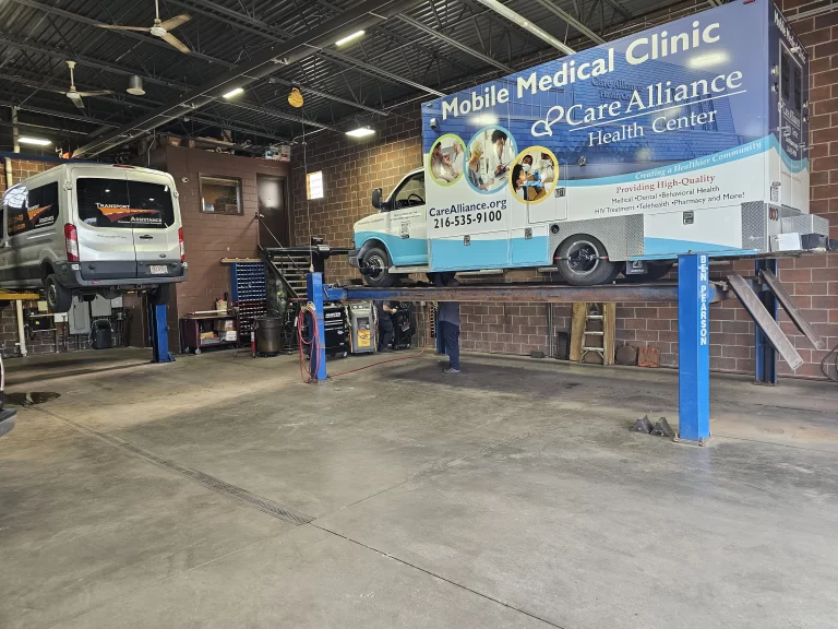 A close-up of a mobile medical clinic truck on a four-post car lift and an SUV on an Overhead Car Lift inside an auto repair shop garage.
