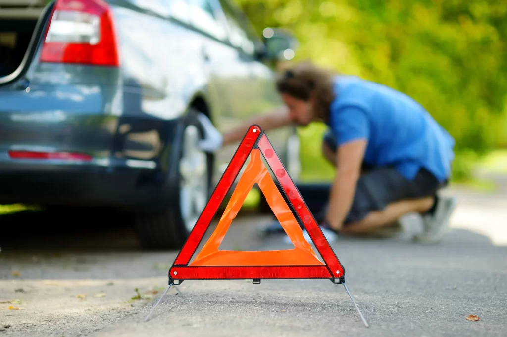A red triangle warning sign on the road with a man and a car parked on the side of the road.